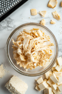 Top-down view of a clean kitchen countertop with a drained can of jackfruit being shredded by hand, seeds and firm pieces removed. Next to it, super firm tofu is being grated using a box grater. The shredded jackfruit and grated tofu are combined in a bowl, ready for cooking.