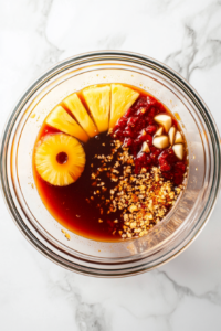 A top-down view of a large mixing bowl on a white marble cooktop. Ingredients like pineapple rings, soy sauce, sesame oil, garlic, ginger, brown sugar, ketchup, tomato paste, red pepper flakes, nutmeg, and chili powder are being whisked together, creating a smooth, colorful marinade