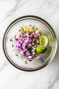 Top-down view of a mixing bowl on a white marble countertop with ⅓ cup of finely diced red onion soaking in ¼ cup of fresh lime juice. The scene captures the bright red onions blending with the lime juice.