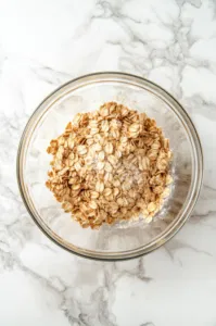 Top-down view of the oat topping ingredients – oats, brown sugar, butter, and flour – being mixed with a pastry blender in a bowl until small chunks form.