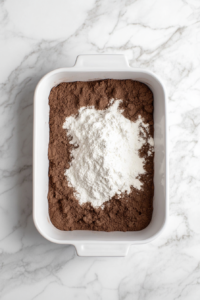 A top-down view of a 9x13-inch ungreased baking dish on a clean kitchen countertop with a white marble cooktop background. The dish contains 3 cups of all-purpose flour, 2 cups of white sugar, 1/2 cup of unsweetened cocoa powder, 2 teaspoons of baking soda, and 1 teaspoon of salt, sifted together to form the base of the cake mixture.