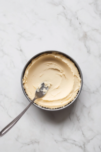 Top-down view of a 23cm springform tin with the biscuit mixture being pressed into the base using the back of a spoon. The base is smooth and even, ready to be chilled in the fridge.