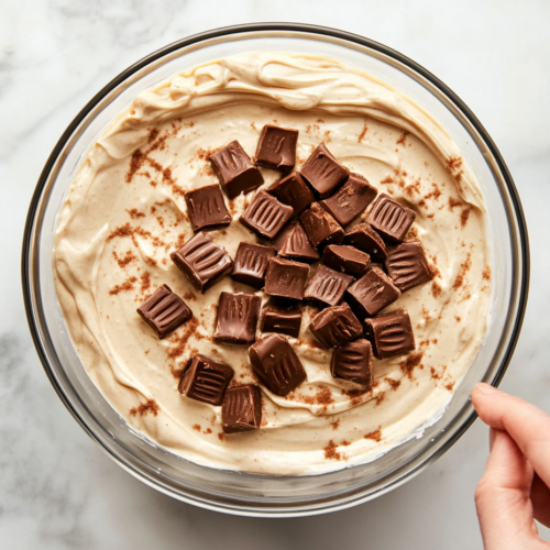 Top-down view of a mixing bowl on a white marble cooktop. Thawed Cool Whip is being folded into the peanut butter mixture with a spatula. Quartered Reese’s Minis are also folded in, creating a light and fluffy dip texture.