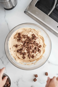 Top-down view of peanut butter cream cheese dip being transferred into a serving bowl on a white marble cooktop. Quartered Reese’s Minis are sprinkled on top for garnish, showcasing the creamy dip ready to be served.