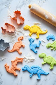 This image shows the colorful dough being rolled out to 7-8mm thickness and dinosaur shapes being cut out with a cookie cutter.