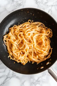 Top-down view of noodles being sautéed in a large skillet with minced ginger and garlic, showing light browning. The skillet is placed on a white marble countertop.