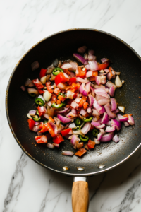 Top-down view of a large pan on a white marble cooktop with avocado oil, sautéing finely diced onion and minced garlic for 5-6 minutes until golden and fragrant.