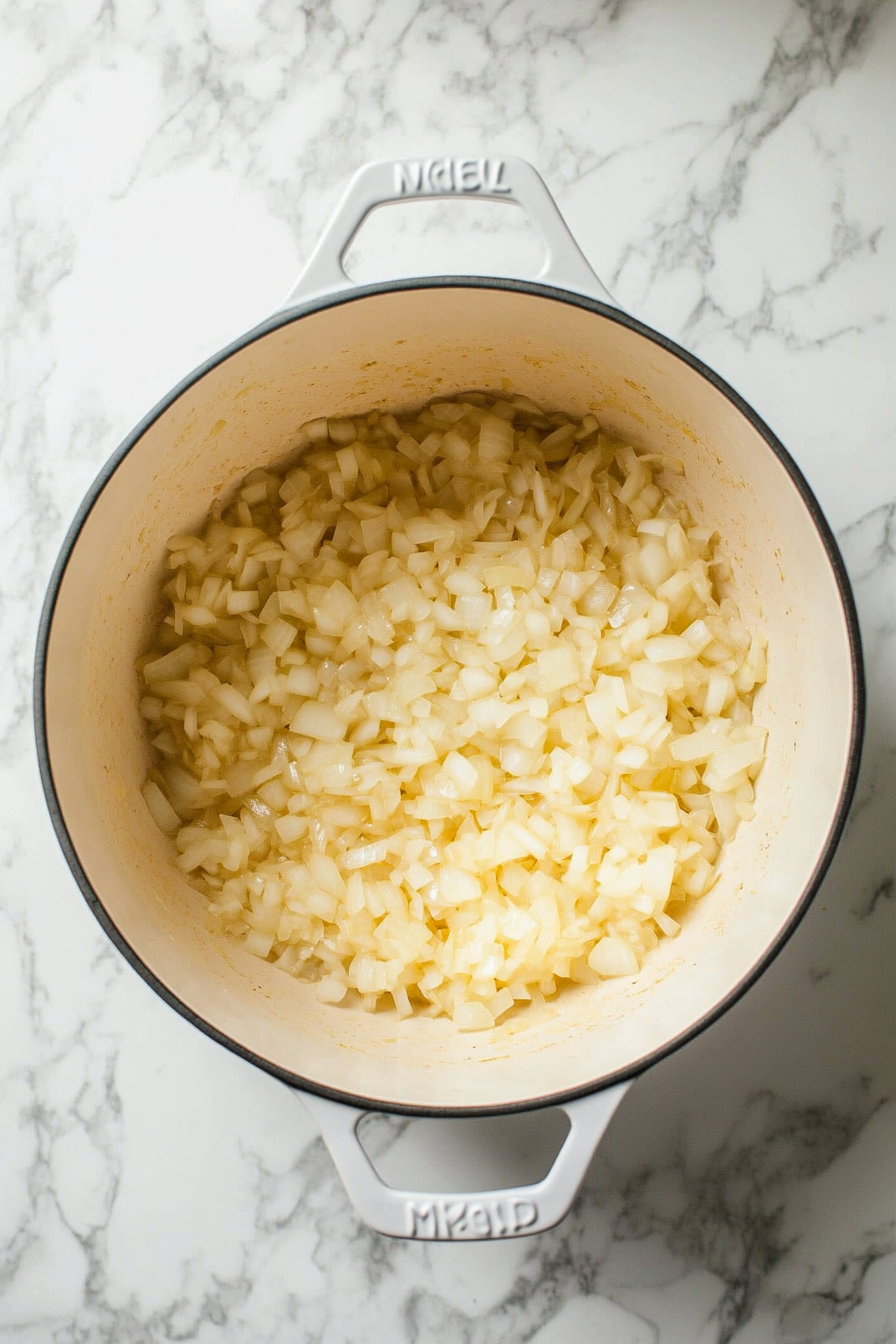 Top-down view of a Dutch oven on a white marble countertop, with ½ chopped yellow onion sautéing with sea salt and a splash of water over medium-low heat. The onions are translucent and gently cooking.