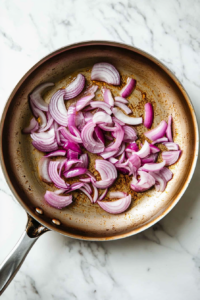 Top-down view of a large pan on a white marble cooktop with avocado oil heating. Thinly sliced red onions are being sautéed until translucent and golden, sizzling in the hot oil for about 3-5 minutes.