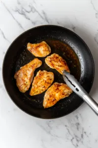 Top-down view of a skillet on a white marble cooktop with olive oil and butter sizzling as chicken pieces are searing to a golden brown. The scene captures the chicken being flipped with tongs to achieve a crispy crust on both sides.