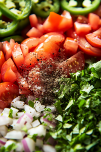 Top-down view of salt and cracked black pepper sprinkled over the tomato, onion, and cilantro mixture. The seasoning is being stirred in, enhancing the fresh salsa flavors.