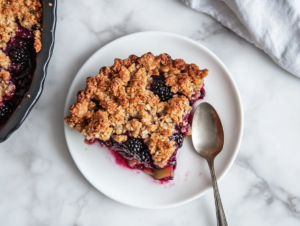 Top-down view of a slice of blackberry rhubarb crumble on a clean white plate on the white marble cooktop. The slice shows the golden, crunchy crumble topping and juicy fruit filling. A spoon is placed next to the serving, highlighting the contrast between the crispy topping and the soft, flavorful fruit.