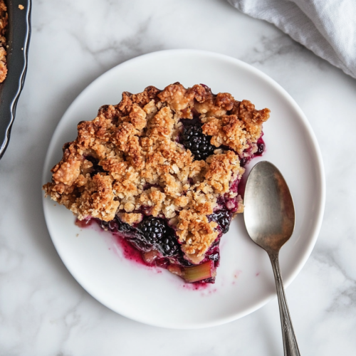 Top-down view of a slice of blackberry rhubarb crumble on a clean white plate on the white marble cooktop. The slice shows the golden, crunchy crumble topping and juicy fruit filling. A spoon is placed next to the serving, highlighting the contrast between the crispy topping and the soft, flavorful fruit.