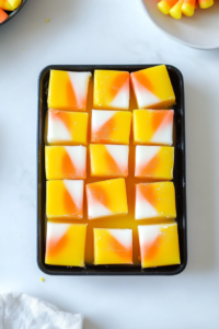 Top-down view of the Candy Corn Fudge after being sliced into small squares, showing its distinct yellow, orange, and white layers stacked atop one another. The fudge is arranged on a white dish, ready to serve on a white countertop.