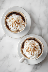 Top-down view of two mugs of Irish coffee on a clean white saucer on the white marble cooktop. The whipped cream is topped with a light dusting of nutmeg, and the hot coffee is ready to be enjoyed, with steam rising from the mugs.