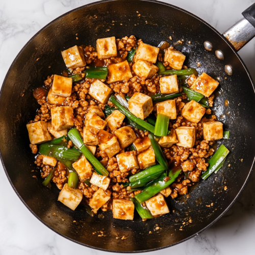 A top-down view of the finished dish in a wok on a white marble cooktop background. The tofu, ground pork (or chicken), and scallions are fully combined, with the sauce coating everything evenly, ready to be served.