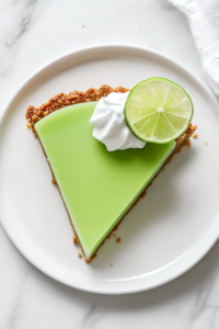 Top-down view of a slice of lime Jello pie on a white plate. The slice reveals the creamy filling in the graham cracker crust, topped with whipped topping and a thin slice of lime, ready to be enjoyed.