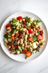 A top-down view of a serving of bean and cucumber salad on a clean white plate on a white marble cooktop. The colorful mixture of beans, cucumber, capers, and parsley is coated with the tangy dressing, ready to be enjoyed.