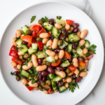 A top-down view of a serving of bean and cucumber salad on a clean white plate on a white marble cooktop. The colorful mixture of beans, cucumber, capers, and parsley is coated with the tangy dressing, ready to be enjoyed.