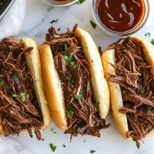 This image shows a close-up of a tender, juicy BBQ beef sandwich, filled with shredded beef and topped with barbecue sauce, served on a soft toasted bun with coleslaw on the side.