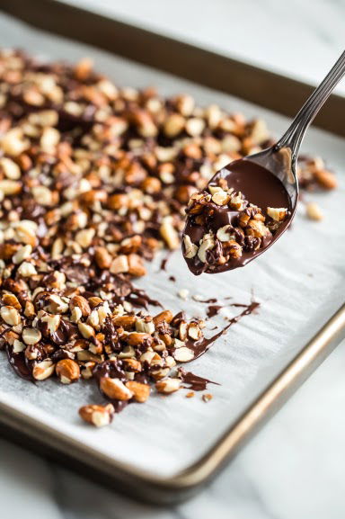 Top-down view of a wax paper-lined baking sheet on a white marble cooktop. Small spoonfuls of the chocolate peanut mixture are dropped onto the sheet, forming individual glossy peanut clusters, evenly spaced and ready to chill.