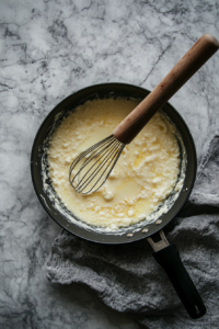 Top-down view of a saucepan with 2 tablespoons of flour being whisked into a butter and garlic mixture, forming a smooth roux. The white marble cooktop surrounds the scene as the mixture thickens.