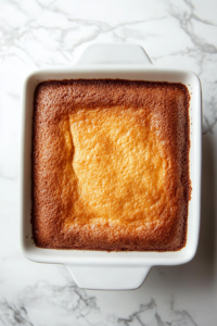 A top-down view of the baked cake in the 9x13-inch dish on a white marble countertop. A toothpick is being inserted into the center of the cake to test for doneness. The toothpick comes out clean, indicating the cake is fully baked and ready to be frosted.