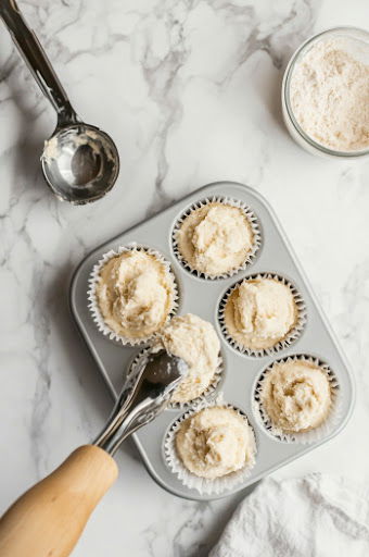 This image shows a medium-sized cookie scoop being used to portion the batter into a cupcake pan lined with paper liners.