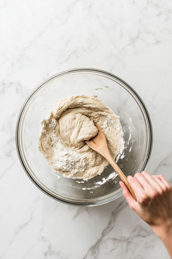 This image shows flour, yeast, and salt being mixed together in a large bowl with a wooden spoon.