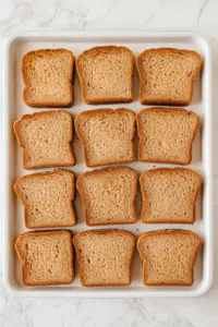 This image shows slices of bread arranged in a single layer on two baking sheets, ready to be toasted in the oven until dry.