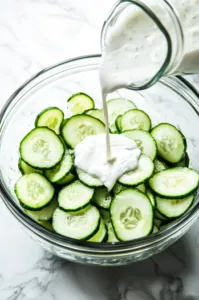 Top-down view of sour cream dressing being poured over cucumber slices in a large bowl, with the salad being gently tossed to evenly coat all the slices.