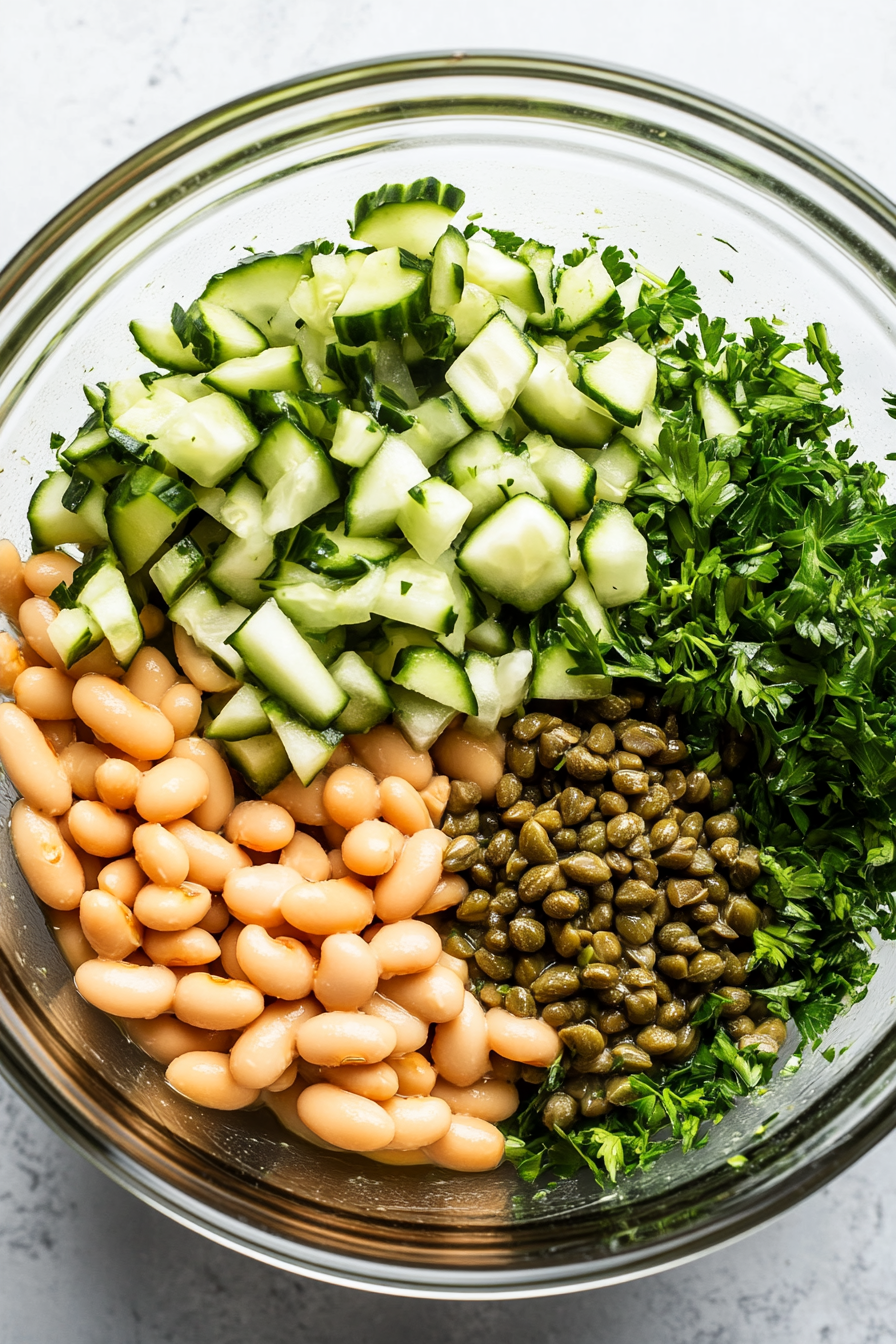 A top-down view of a large mixing bowl filled with drained onions, beans, cucumber, capers, parsley, and oregano being tossed together with a dressing. The vibrant colors of the ingredients stand out in the salad preparation.