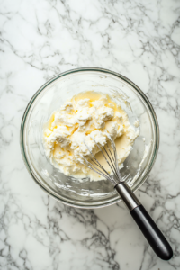 This image shows a hand-held electric mixer whipping together heavy cream, softened cream cheese, lemon juice, and confectioners' sugar in a bowl. The creamy lemon filling is being whipped to a fluffy consistency with hints of vanilla and fresh lemon zest to give it a smooth, tangy flavor.