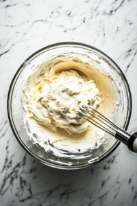 A top-down view of a mixing bowl on the white marble cooktop. Heavy whipping cream is being whipped with an electric mixer until stiff peaks form. The whipped cream is light and airy, ready to be chilled. The scene focuses on the texture of the whipped cream as it reaches the perfect consistency.