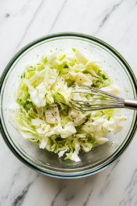 Top-down view of a mixing bowl on a white marble cooktop with olive oil, red wine vinegar, celery seed, and sugar being whisked together to create the dressing.