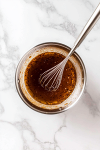 A top-down view of a small saucepan on a clean kitchen countertop with a white marble cooktop background. Inside the saucepan, hoisin sauce, soy sauce, honey, and Chinese five-spice powder are being whisked together. The mixture is smooth and glossy, with a whisk stirring the ingredients. The scene focuses on blending the sauces and spices.