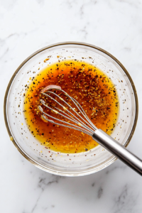 A top-down view of a large mixing bowl on a white marble cooktop, with ingredients like red wine vinegar, olive oil, Dijon mustard, sea salt, and black pepper being whisked together to create a smooth, tangy dressing.