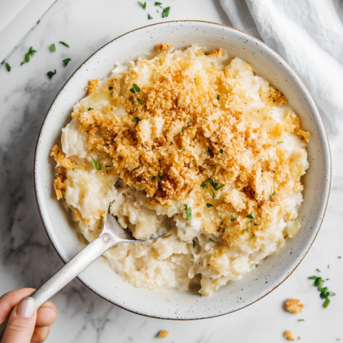 A top-down view of the finished dish being served from the slow cooker onto a plate on a clean kitchen countertop with a white marble cooktop background. The creamy, cheesy hash brown mixture with a crunchy cracker topping is being spooned onto the plate, ready to be enjoyed. The scene focuses on the rich texture and golden topping of the dish, with the plate and serving spoon clearly visible.