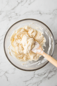 Top-down view of vanilla extract being added to creamed butter and sugar in a bowl, with flour and salt gently mixed in with a spatula on a white marble cooktop.