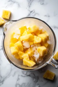 Top-down view of frozen pineapple chunks and ice cubes being added to the pitcher, on a bright surface with a summery vibe.