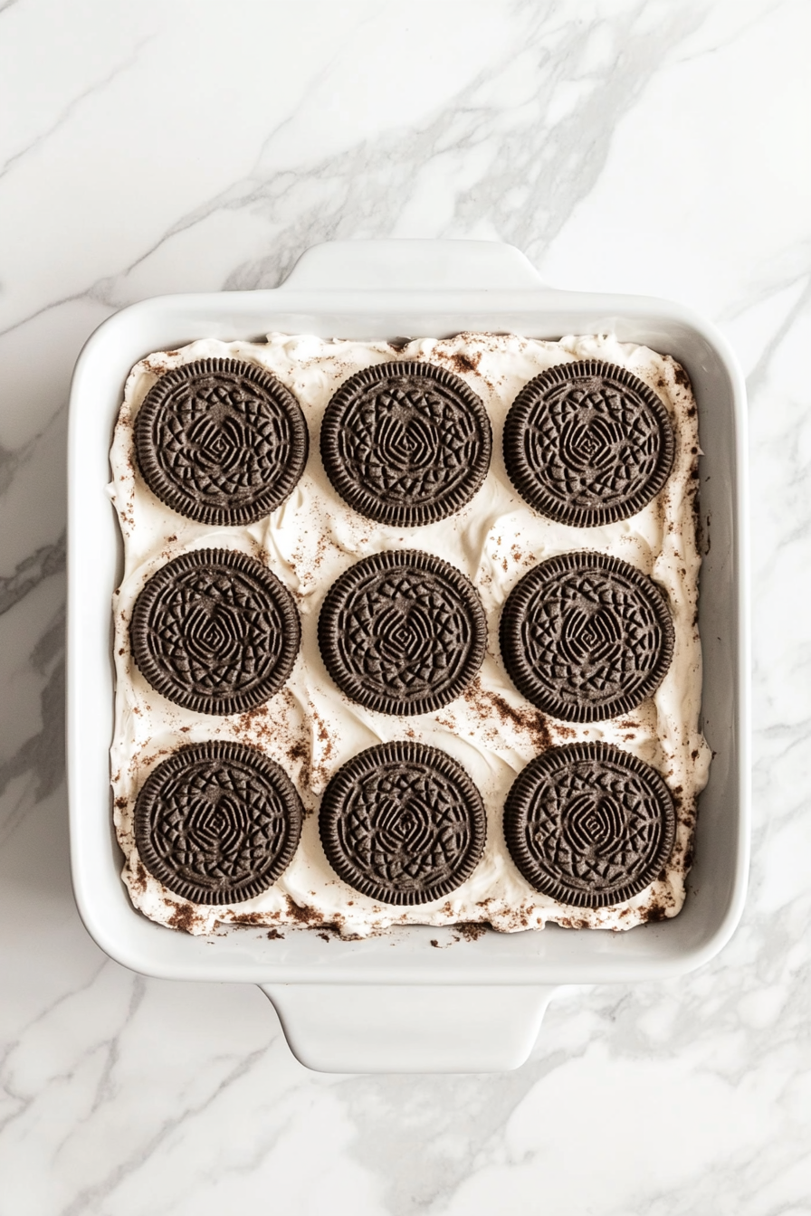 Top-down view of a baking dish with a cookie dough layer on a white marble countertop.Oreos are arranged neatly in four rows on top of the cookie dough, evenly spaced.