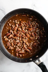 Top-down view of chopped pecans being stirred into the hot toffee mixture in the saucepan, combining well.