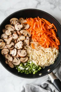 Top-down view of sliced mushrooms, water chestnuts, blanched beans, and carrots being added to the skillet with sautéed noodles. The vibrant vegetables are sizzling in the skillet on a white marble countertop.