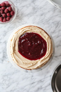 Top-down view of a cake being assembled with peanut butter frosting and raspberry jam layered between the peanut butter cake layers.