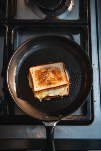 Top-down view of a skillet with a sandwich cooking on medium-low heat. The bread is toasting and cheese is beginning to melt.