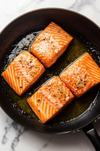 Top-down view of salmon fillets, skin side down, sizzling in a large nonstick skillet with olive oil on a white marble cooktop. The skin is starting to crisp.