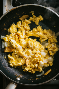 Top-down view of a skillet with a thin layer of oil left after draining. Fried tortillas and onions are returned to the skillet with 6 lightly beaten eggs being added. The mixture is being stirred frequently as the eggs cook and set, showing crispy tortillas and scrambled eggs seasoned with salt.