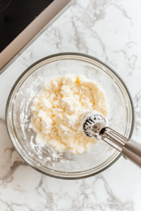 Top-down view of butter and confectioners' sugar being creamed together in a mixing bowl with an electric mixer on a white marble cooktop.