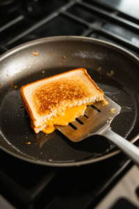 Top-down view of a skillet with a sandwich being flipped with a spatula, showing a golden and toasted underside with melting cheese.