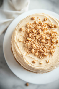 Top-down view of a fully frosted peanut butter cake being garnished with chopped peanuts, adding texture and visual appeal.